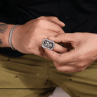 Man adjusting a silver flip ring with ornate double initials on his finger, set against a casual attire background.