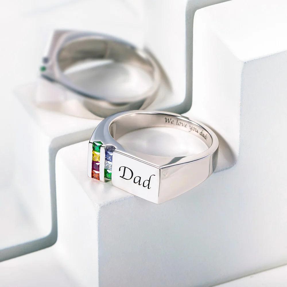 Silver ring with "Dad" and colorful gemstones displayed on a reflective surface, with a mirrored ring in the background.