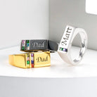 Assortment of rings displayed: one black and one gold engraved with "Dad," and one silver with "Matt," all adorned with gemstones.