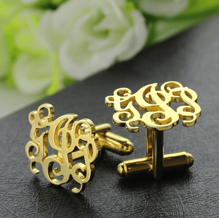 Elegant gold monogram cufflinks with script initials, displayed on a leather surface with white roses in the background.
