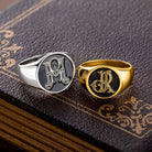 Silver and gold tone custom monogram signet rings with intertwined initials displayed on a decorative book.