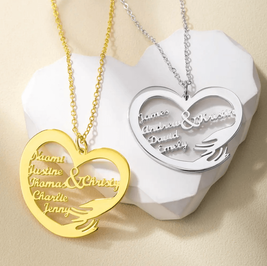 Two heart-shaped necklaces, one gold and one silver, each engraved with different names, displayed on a light-colored surface with a white heart-shaped ornament.