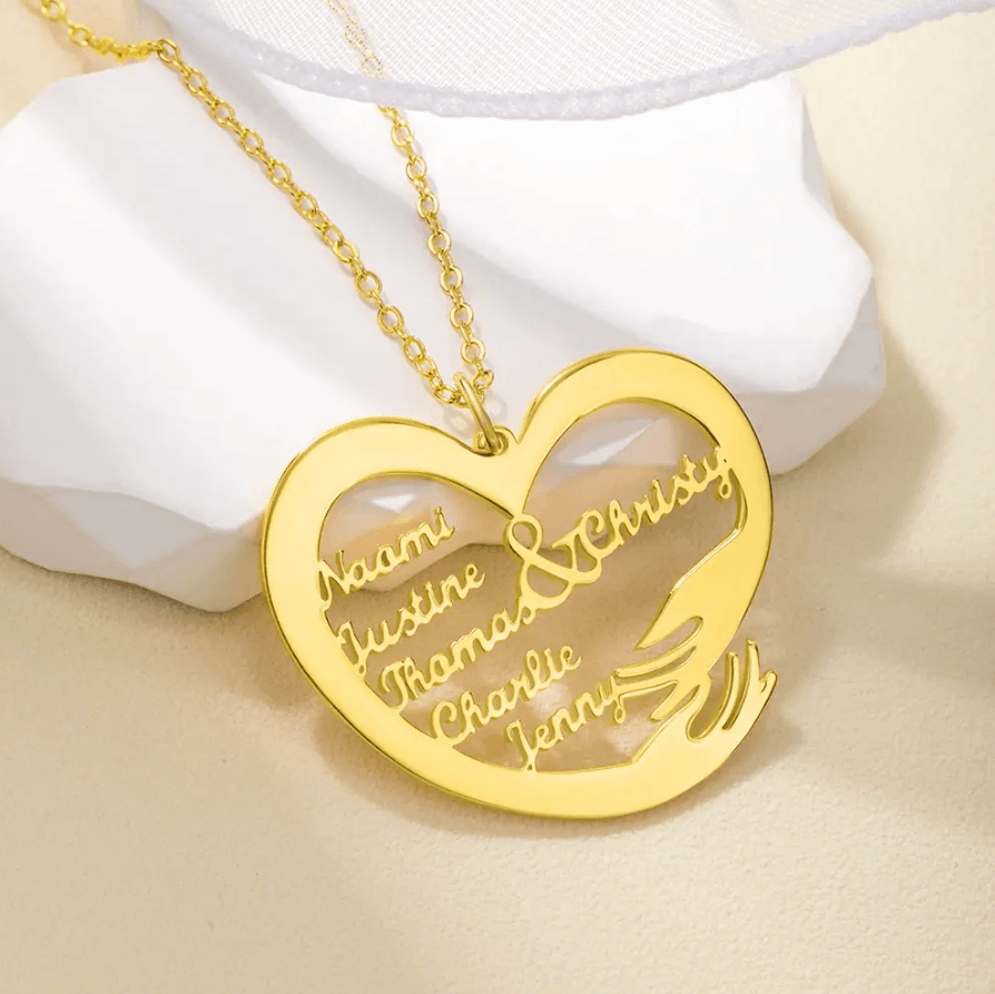 A heart-shaped gold necklace pendant engraved with the names "Naomi, Justine, Thomas, Christy, Charlie, Jenny," displayed on a light-colored surface with a white ornament.