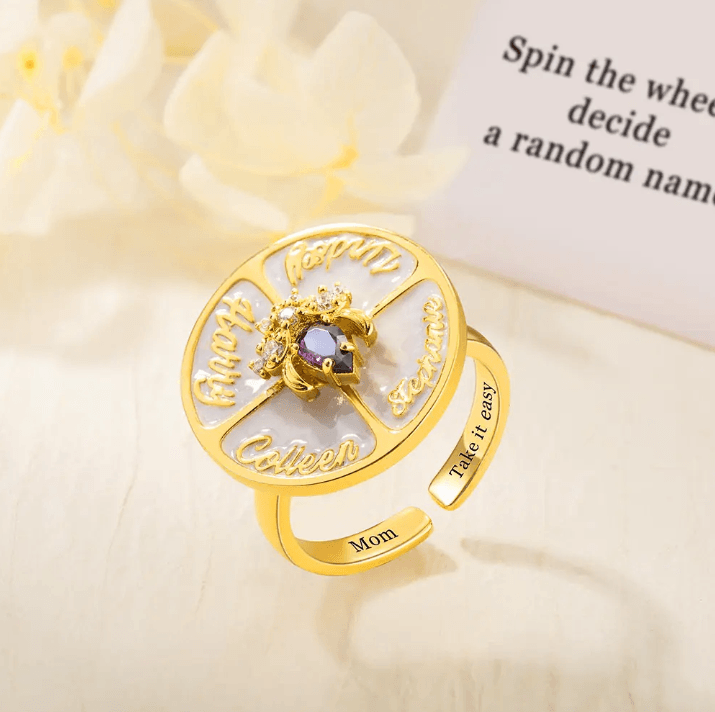 Gold spinner ring with engraved family names and a central birthstone, used for making fun decisions and as a customizable, unique gift.