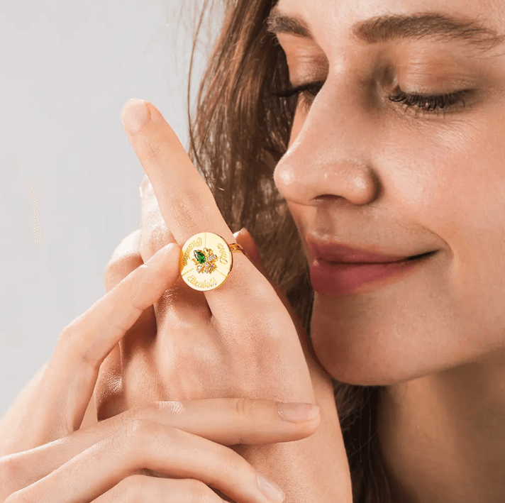 Smiling woman wearing a gold spinner ring with engraved names and a central green birthstone, showcasing the ring's unique and customizable design.