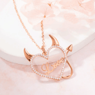 Rose gold necklace with a heart-shaped pendant featuring devil horns and tail, and the word "Love" in the center, surrounded by sparkling stones, displayed on a soft pink surface.