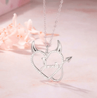 Silver necklace with a heart-shaped pendant featuring devil horns and tail, and the name "Jennie" in the center, surrounded by sparkling stones, displayed on a soft pink surface.