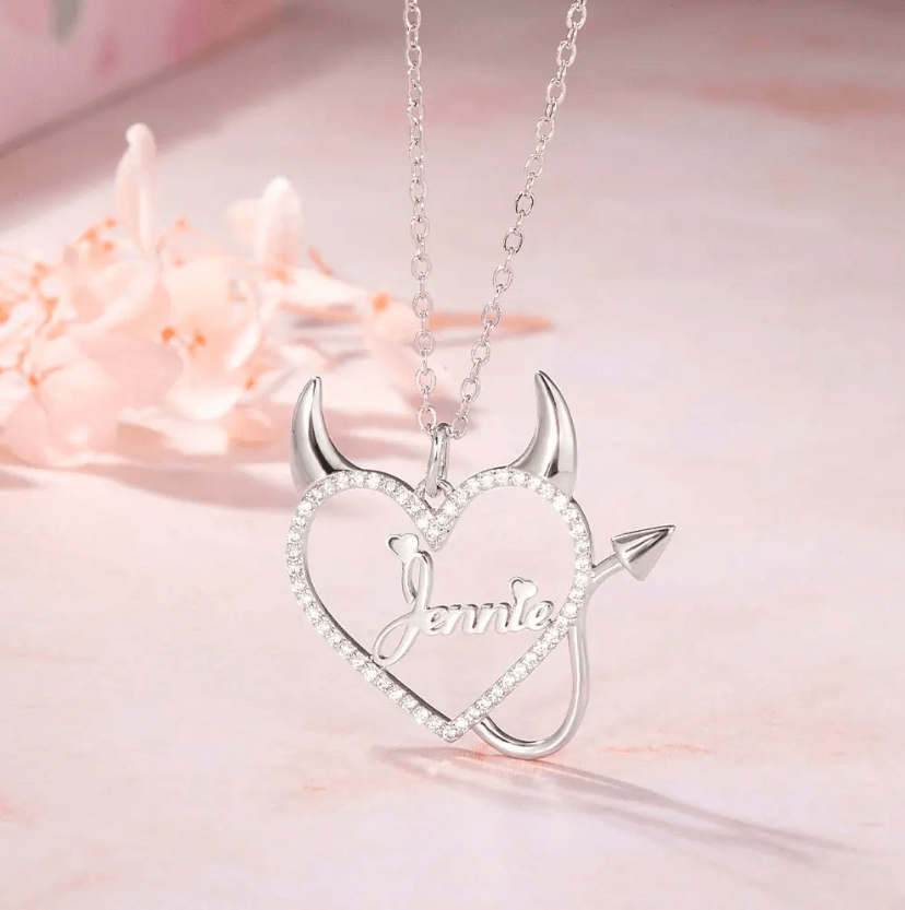 Silver necklace with a heart-shaped pendant featuring devil horns and tail, and the name "Jennie" in the center, surrounded by sparkling stones, displayed on a soft pink surface.