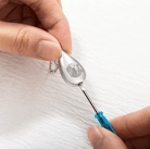 Hand assembling a silver teardrop urn necklace with an engraved dog's face, using a blue-handled screwdriver.