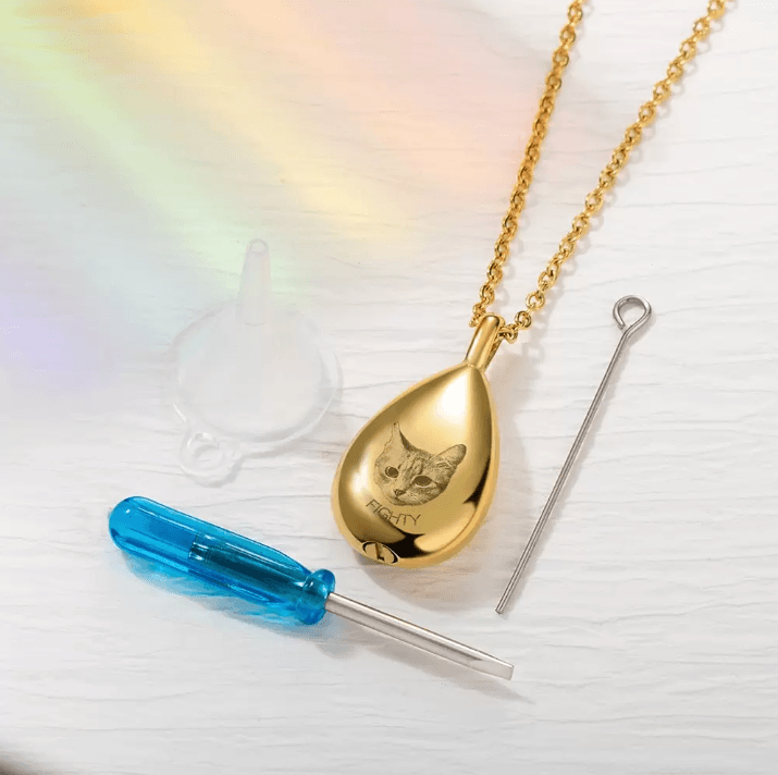 Gold teardrop urn necklace with cat engraving, alongside tools for assembly on a rainbow-tinged background.