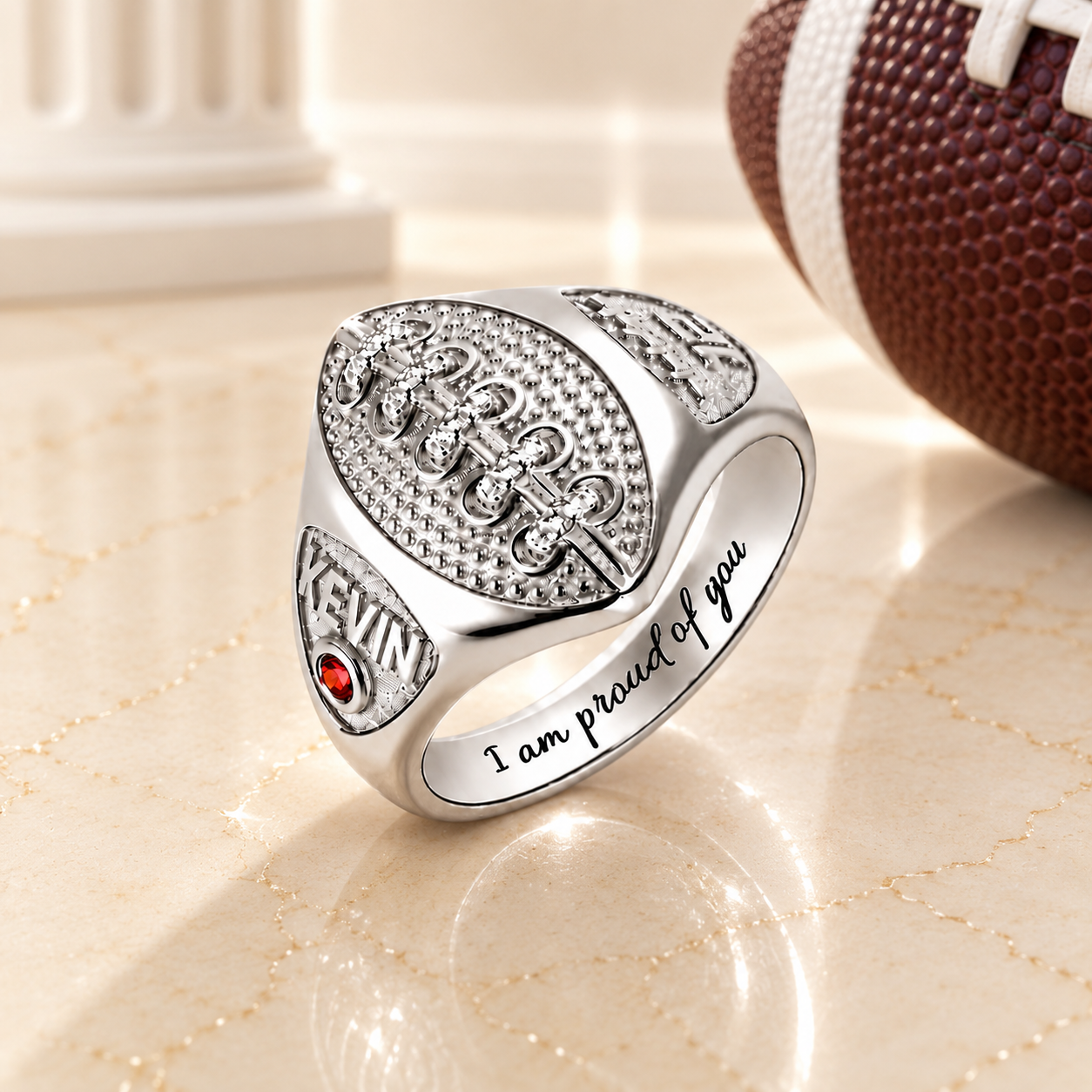 Personalized silver football ring with red birthstone, engraved name Kevin, and “I am proud of you” message on a black marble surface.