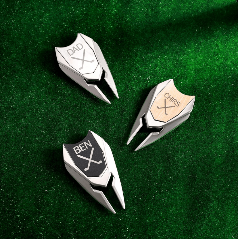 Three personalized stainless steel golf marker divot repair tools with custom monograms "DAD," "CHRIS," and "BEN," displayed on green turf.