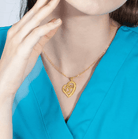 Gold heart-shaped pendant with stethoscope design and letter 'D' charm, on a woman in a teal blouse.