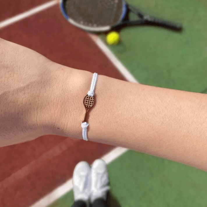 A wrist wearing a white cord bracelet with a small gold tennis racket charm, with a tennis court, racket, and ball in the background.