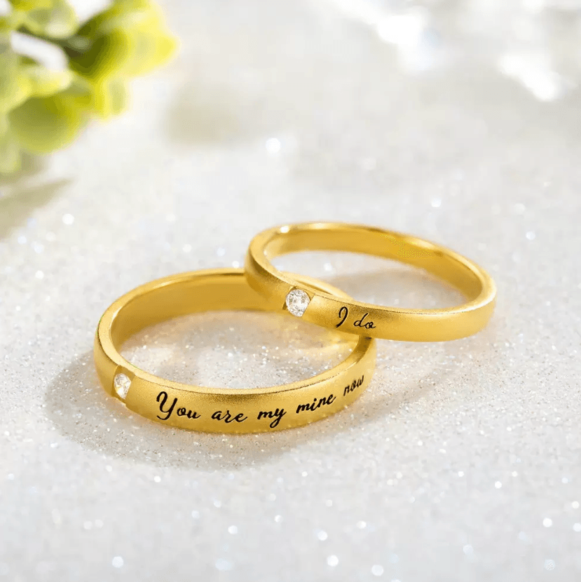 Two golden rings on a glittery surface, one engraved with 'I do' and the other 'You are my mine now', both with diamonds.