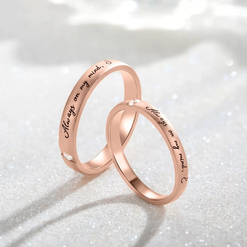 Two rose gold rings intertwined, engraved with 'Always on my mind' and 'Forever in my heart', set against a glittery background.