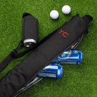 An insulated black golf beer can bag with a customizable red monogram lies on a green artificial turf next to two golf balls. The bag has an open compartment revealing a stack of cold beer cans inside, and an attached side koozie with a can halfway inserted. The bag features a shoulder strap for easy carrying.