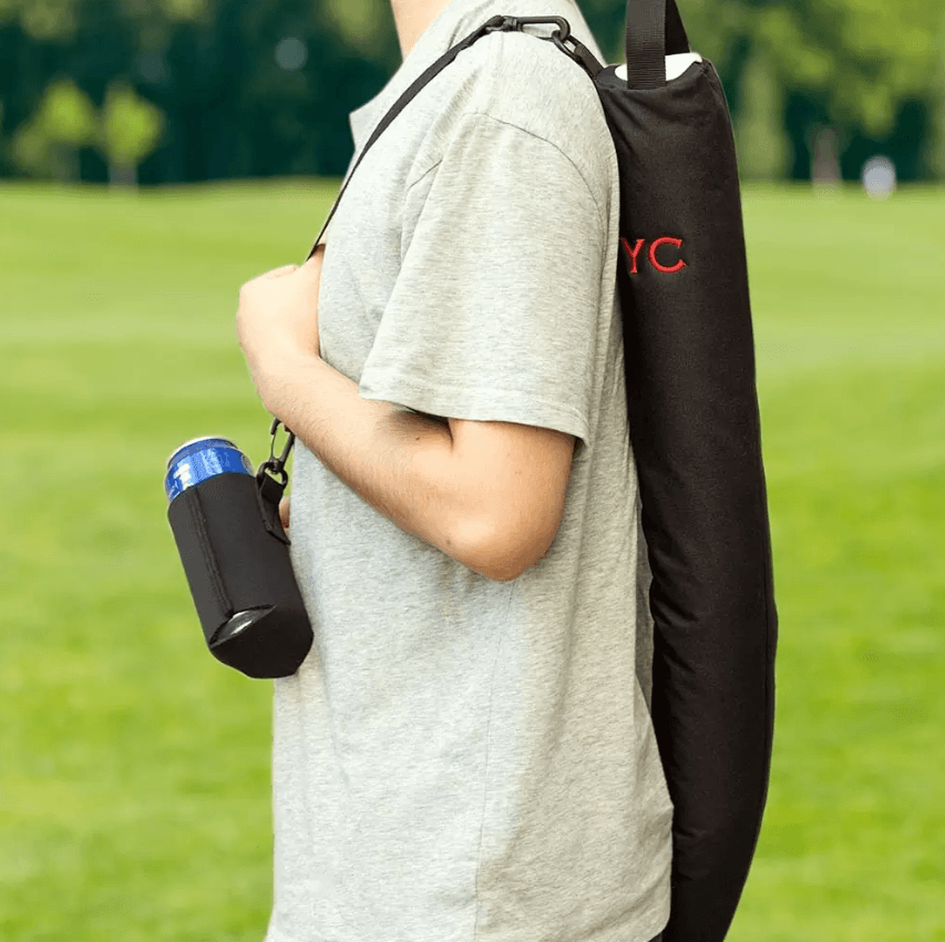 A person, visible from the shoulders down, stands on a grassy area with a black insulated beer can bag slung over one shoulder, featuring the initials 'YC' in red. The individual is also holding a can of beer in a black koozie, ready for enjoyment. The scene suggests a casual, leisurely atmosphere, likely on a golf course.