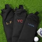 Three black insulated beer can bags with personalized embroidery lying on green artificial turf. From left to right, the first bag has 'L.F.' embroidered in gold, the middle bag has 'YC' in red, and the third bag has 'LEON' in blue. Next to them is a white golf ball and a can of beer, suggesting these bags are designed for carrying beverages on the golf course. The bags have loops with carabiners for easy attachment.