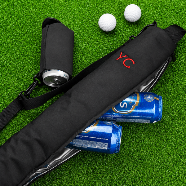 Personalized black can cooler with red YC initials, holding beer cans on green turf with golf balls nearby