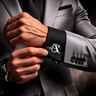 Silver interlocking initials cufflink worn on a black shirt cuff with a grey suit.