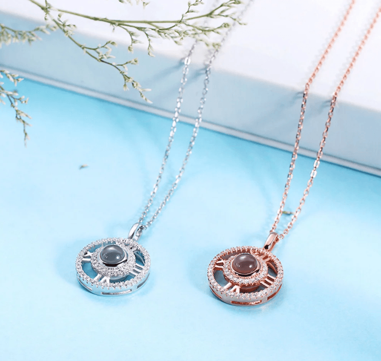 Two elegant necklaces with circular pendants featuring Roman numerals and central gemstones, one in silver and the other in rose gold, displayed on a light blue surface.