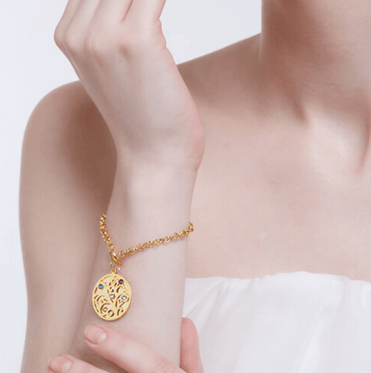 A woman in a white strapless top wearing a gold charm bracelet with a tree pendant featuring colored gemstones, lightly touching her face with her fingers.