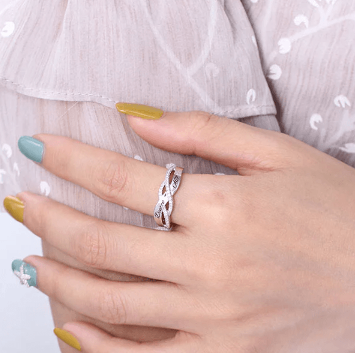 A hand with multi-colored nails wearing a silver infinity ring with diamond accents, engraved with the names "Denton" and "Alex," against a light fabric background.