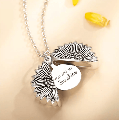 Silver sunflower necklace opened to reveal 'You Are My Sunshine' message, with yellow petals nearby.