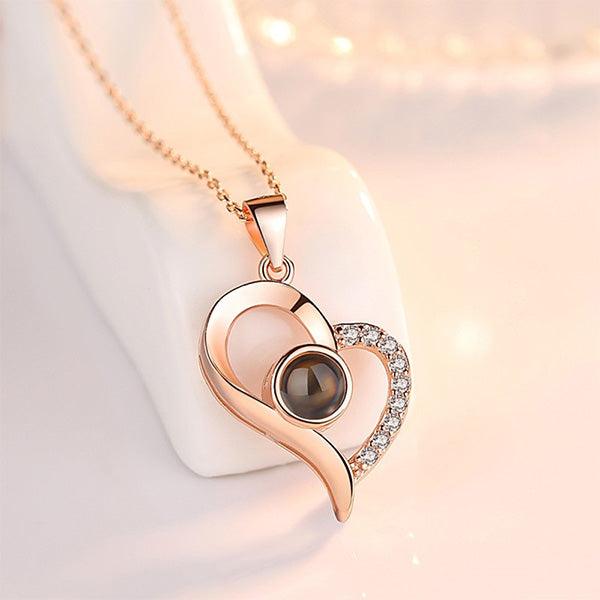 Rose gold heart-shaped pendant necklace with a central black gemstone and small clear crystals on one side, displayed against a soft pink background.