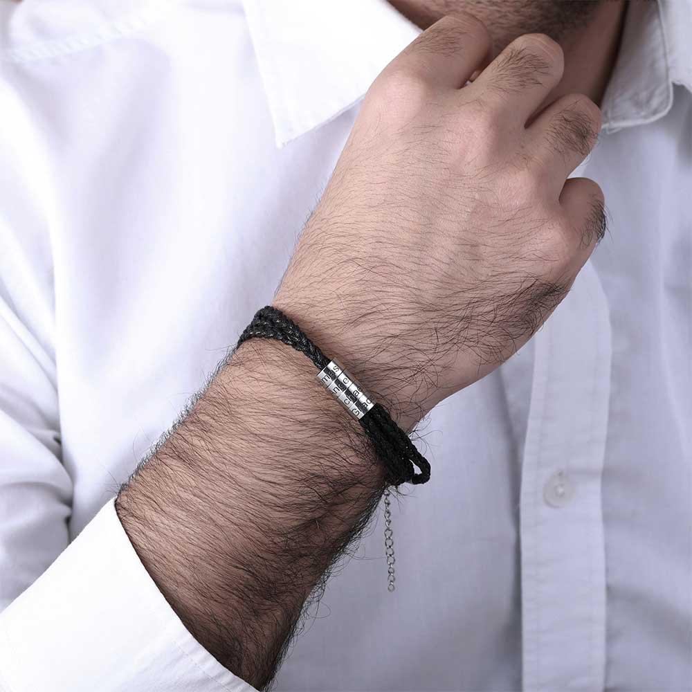 Man wearing a black braided bracelet with silver beads engraved 'Thor', 'Angel', 'Michael', 'Grace', on his wrist.