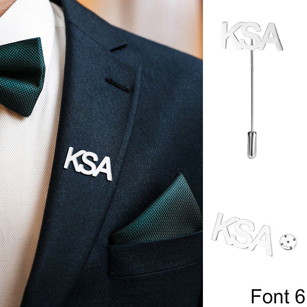 Suit with 'KSA' silver monogram lapel pin, white shirt, and teal tie and pocket square.