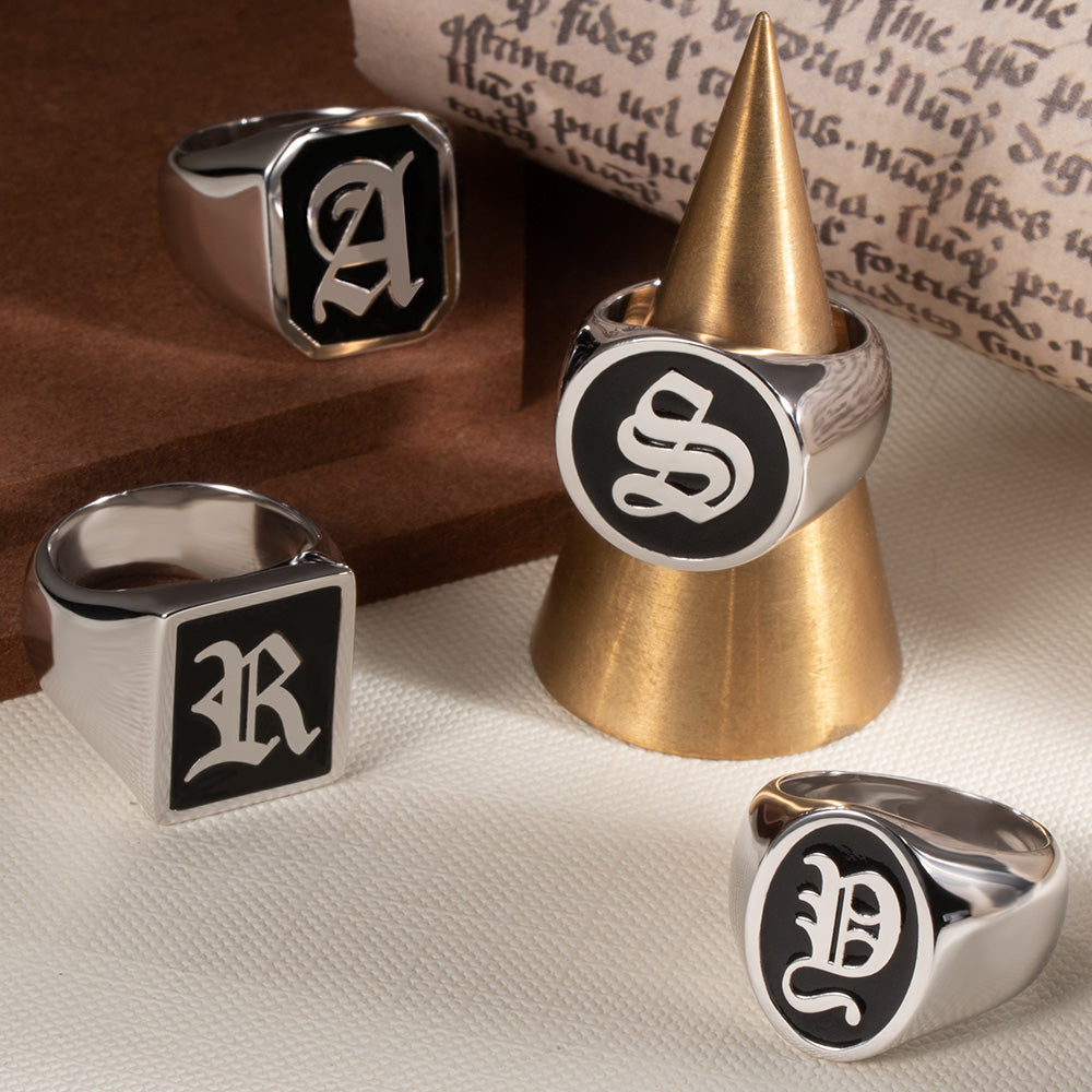 Sterling silver signet rings with black enamel, showcasing initials 'A', 'S', 'R', and an intertwined 'E' and 'L', arranged around a golden cone on a textured cream surface with vintage handwriting as backdrop.