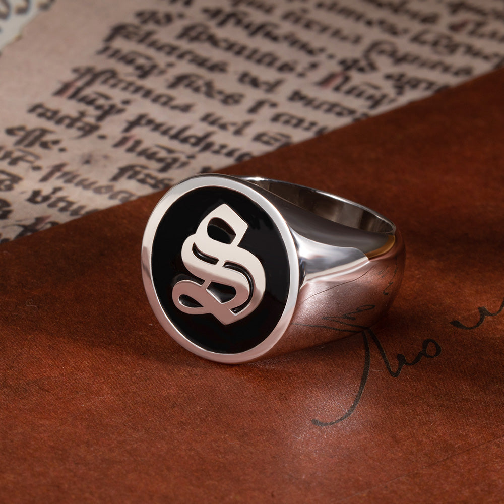 A sterling silver signet ring with a black face and a stylized old English initial "S" in silver, resting on a leather surface with vintage handwriting in the background.