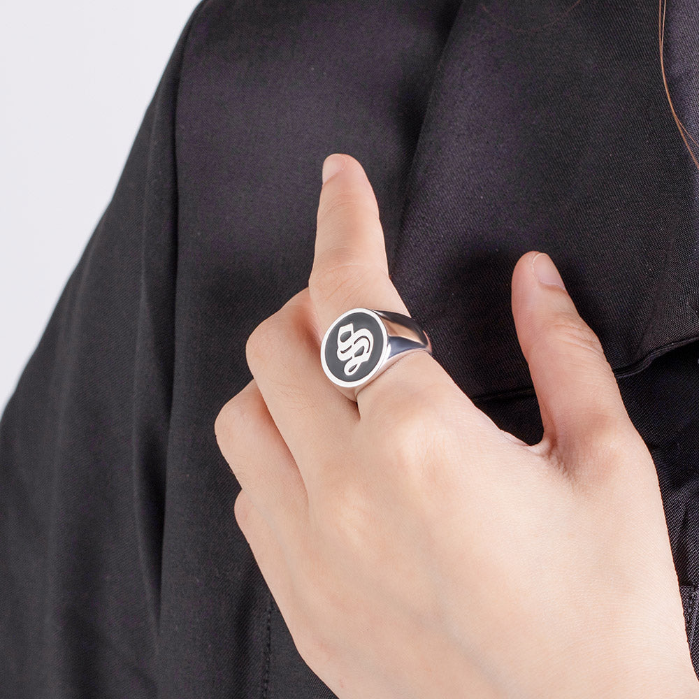 A person's hand against a black fabric background, wearing a sterling silver signet ring with a black enamel face featuring an old English script initial 'S'.