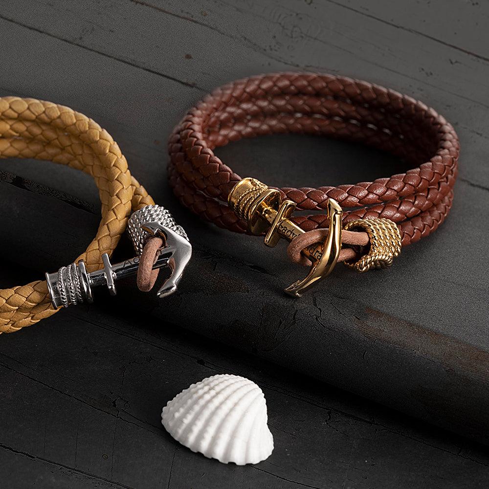 Yellow and brown braided bracelets with silver and gold anchors next to a shell on dark wood.