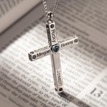 Close-up of a personalized heart cross necklace on a chain, featuring names and birthstones, symbolizing faith and love, placed on an open Bible page.