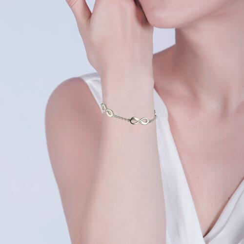 A woman wearing a gold infinity bracelet with a chain link design. The bracelet rests on her wrist as she touches her face, showcasing its elegant style.