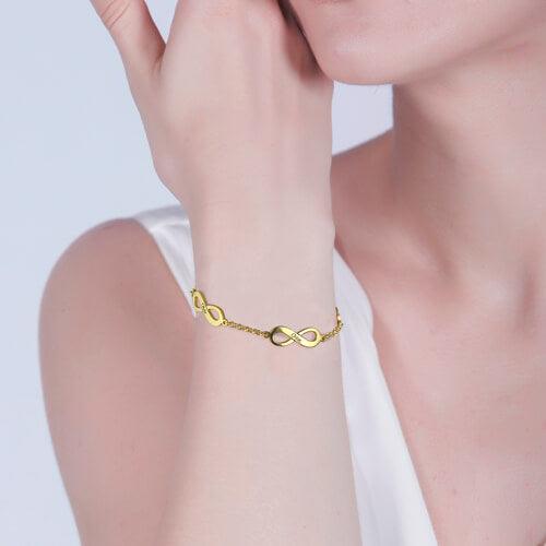 A woman wearing a gold infinity bracelet with a chain link design. The bracelet rests elegantly on her wrist as she lightly touches her face.