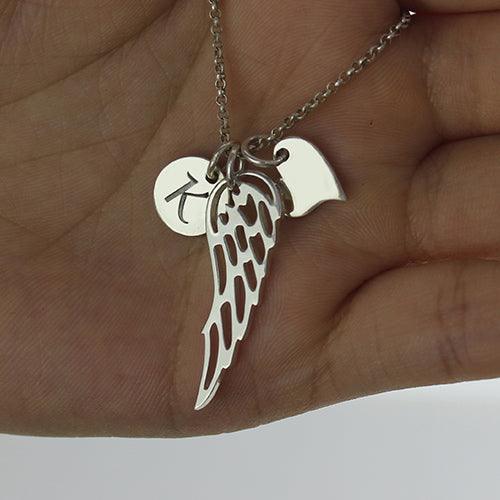 A hand holding a silver necklace with three charms: a circular disc with the letter "K," an angel wing, and a small rectangular tag.