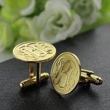 A pair of gold monogrammed cufflinks on a black surface with white roses in the background.
