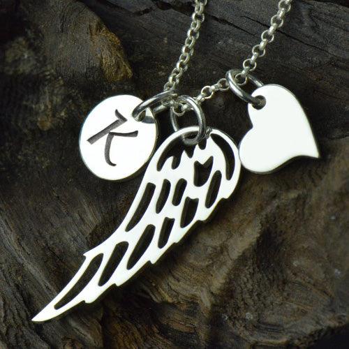A silver necklace with three charms: a circular disc with the letter "K," an angel wing, and a heart, displayed on a dark, textured background.
