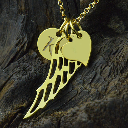 A gold necklace with three charms: a circular disc with the letter "K," an angel wing, and a heart, displayed on a dark, textured background.