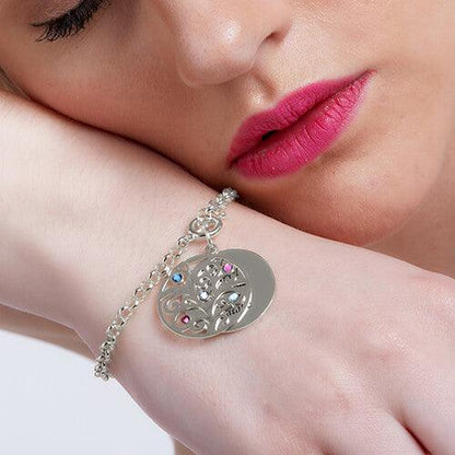 A woman wearing a silver charm bracelet with a tree pendant adorned with colored gemstones and a round disc, resting her head on her arm, with pink lips.