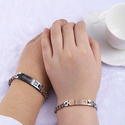 Two hands clasped on a table, each wearing a stainless steel bracelet, beside a white tea cup.