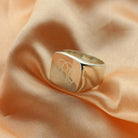 A square silver signet ring with a polished finish, featuring the 1864 Gilles Le Corre monogram style engraved on the face, displaying the initials "ABC," placed on a peach-colored fabric.