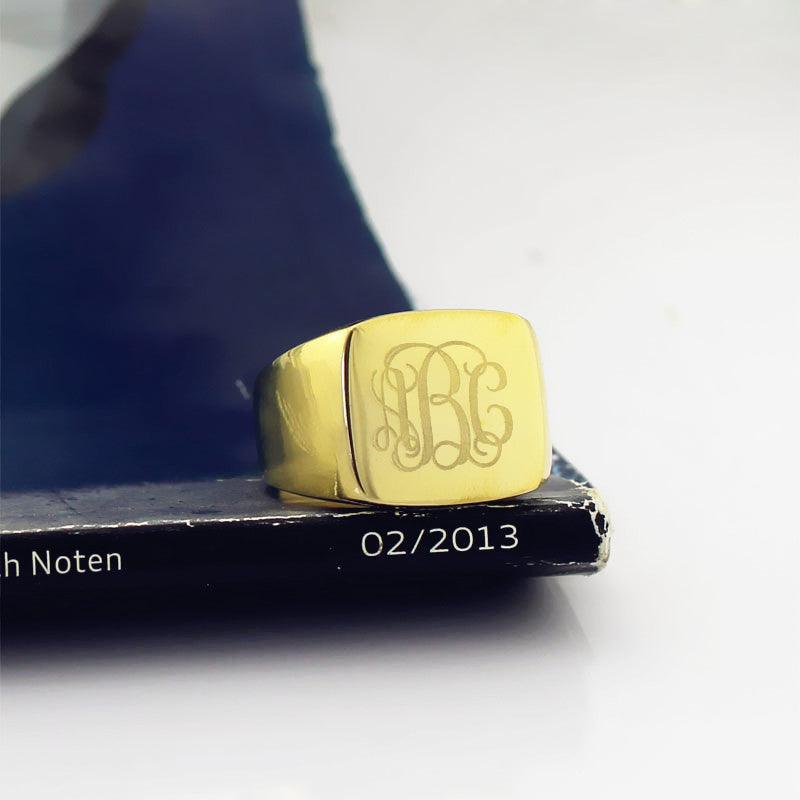 A square gold signet ring with a polished finish, featuring the 1864 Gilles Le Corre monogram style engraved on the face, displaying the initials "ABC," placed on a book with the date "02/2013" visible.