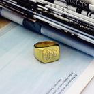 A square gold signet ring with a polished finish, featuring the 1864 Gilles Le Corre monogram style engraved on the face, displaying the initials "ABC," placed on an open book with rolled-up pages in the background.