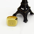 A square gold signet ring with a polished finish, featuring the 1864 Gilles Le Corre monogram style engraved on the face, displaying the initials "ABC," placed next to a small Eiffel Tower model.