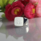 A square silver signet ring with a polished finish, featuring the 1864 Gilles Le Corre monogram style engraved on the face, displaying the initials "ABC," placed next to vibrant pink and red flowers.